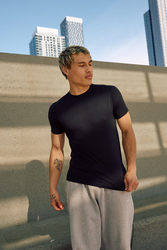 A man wearing a black short-sleeve athletic t-shirt and light gray sweatpants, standing outdoors against a concrete wall with city buildings in the background.