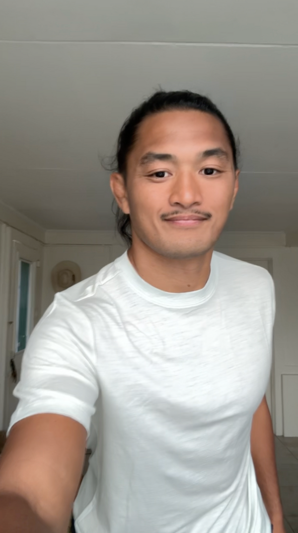 A man with dark hair tied back, wearing a white crew-neck t-shirt, taking a selfie in an indoor room with white walls and ceiling.