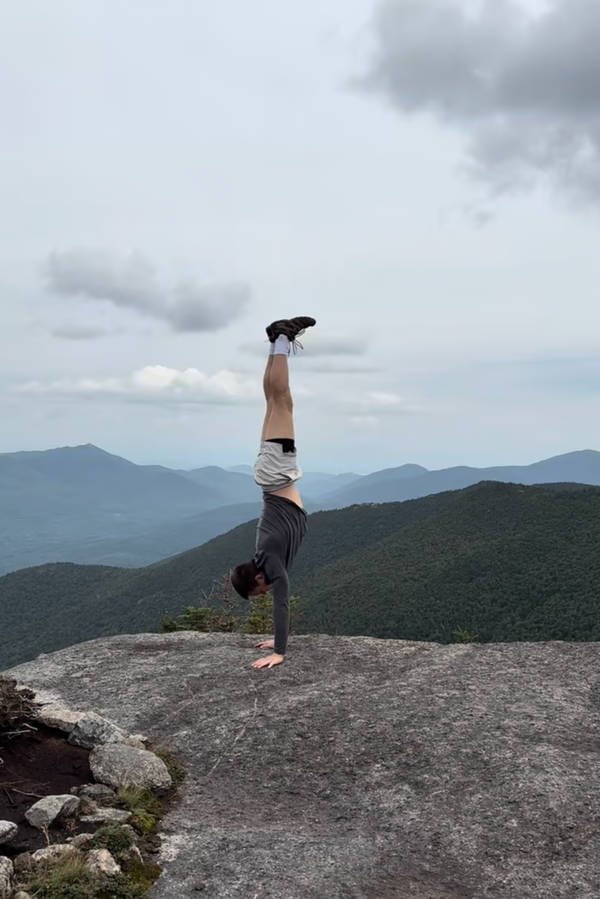 A person performing a handstand on a rocky mountain peak with layered green hills and a cloudy sky in the background.