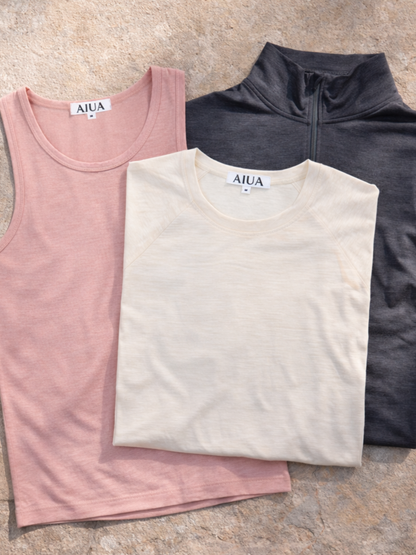 Three folded shirts in pink, beige, and black on a stone surface with 'AIUA' branding.