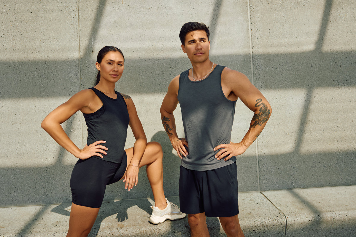 A man and a woman posing in matching athletic wear, including tank tops, shorts, and biker shorts, set against a concrete wall with window shadows.