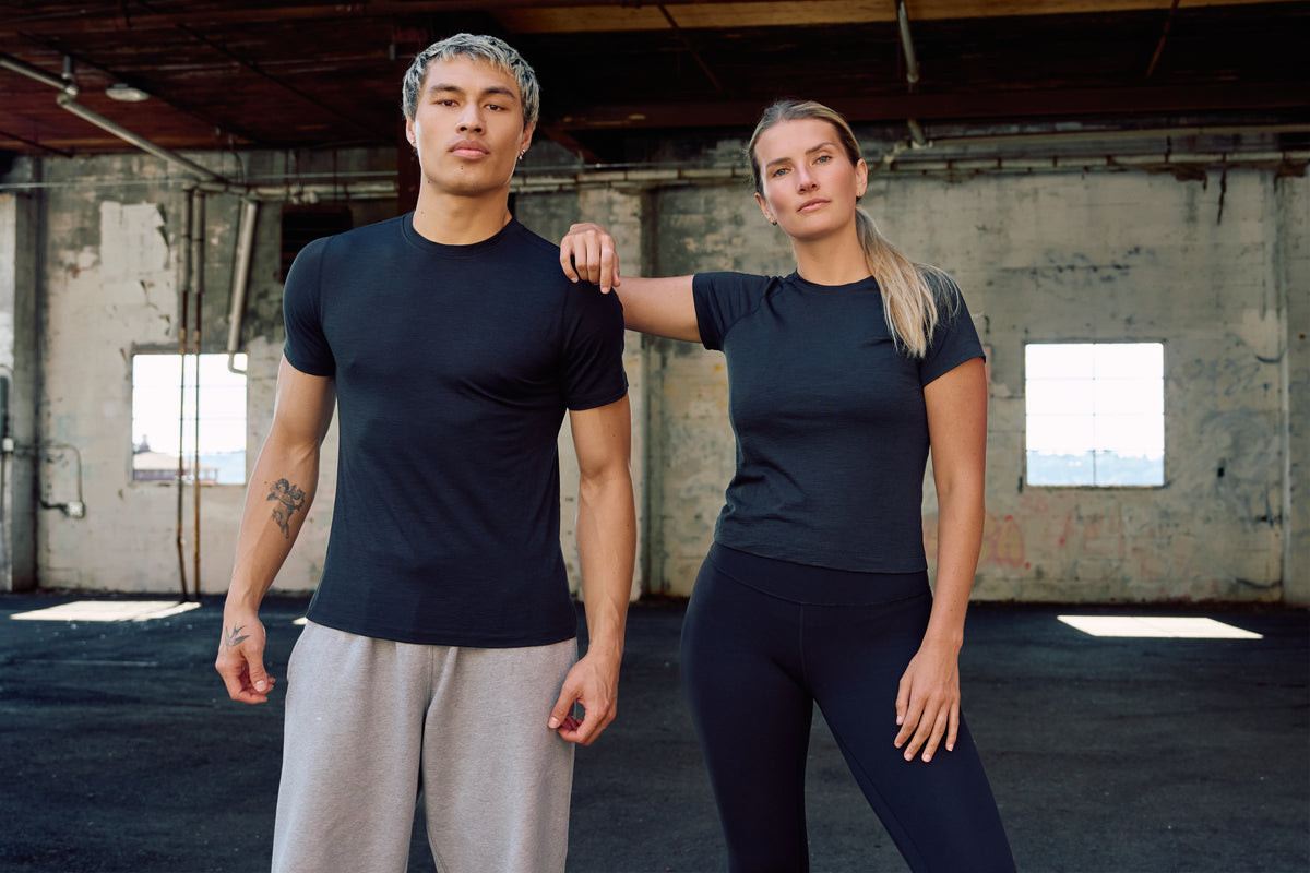 A man and a woman modeling matching dark navy athletic t-shirts in a warehouse setting. The man wears the shirt with light gray sweatpants, and the woman wears the matching shirt with dark leggings.