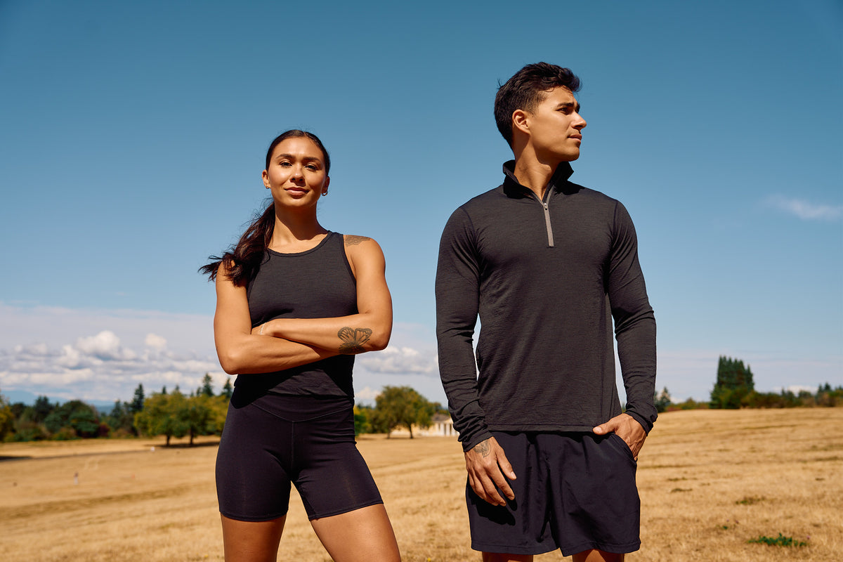 A man and a woman standing outdoors in a dry grass field under a blue sky, wearing dark athletic apparel. The woman wears a black sleeveless tank top and matching biker shorts, while the man wears a black long-sleeve quarter-zip pullover and dark shorts.
