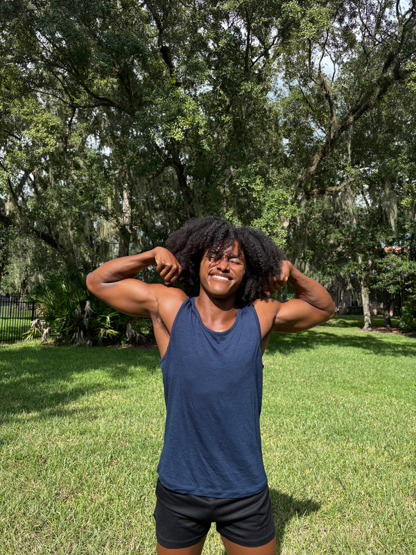 A smiling man stands outdoors on a grassy lawn in front of trees, wearing a navy blue tank top and black athletic shorts, with his arms raised holding his hair.