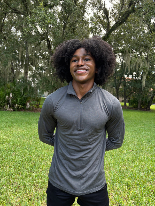 A man outdoors on a grassy lawn in front of trees, wearing a fitted dark gray long-sleeve quarter-zip athletic shirt and black shorts.