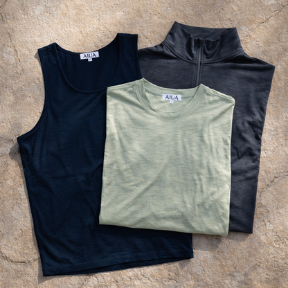 Three folded shirts in black, green, and gray on a stone surface with 'AIUA' branding.