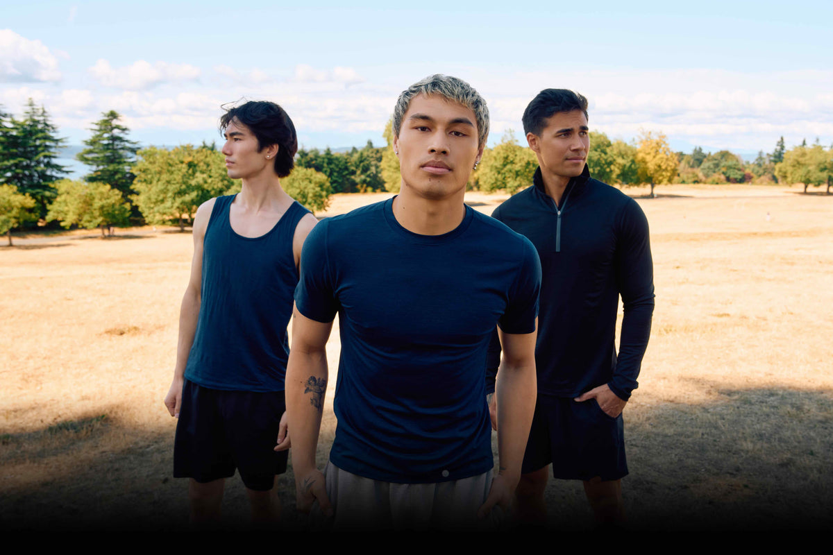 Three men standing outdoors in a dry grass field wearing athletic apparel, including a navy blue crewneck t-shirt in the center, a navy tank top on the left, and a dark navy long-sleeve quarter-zip top on the right.